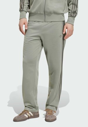 Uomo che indossa pantaloni della tuta Adidas grigio chiaro e giacca abbinata, in piedi con scarpe da ginnastica marroni e bianche visibili.