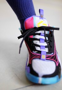 En person iført en flerfarvet atletisk sneaker med sorte snørebånd og en marineblå sok, stående på en lys gulvoverflade.