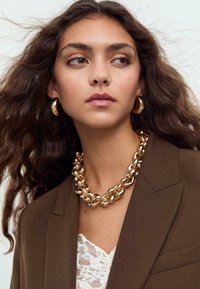 Collier en chaîne en or avec des maillons polis, porté sur un haut en dentelle blanche, associé à des boucles d'oreilles en or et à un blazer marron.