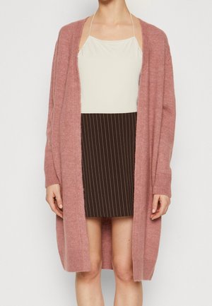 Femme portant un haut blanc à dos nu, une jupe noire à fines rayures et un long cardigan en tricot rose poudré, debout devant un fond uni.