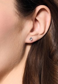 Okrągłe, przezroczyste kryształowe kolczyki na metalowym zapięciu, pokazane na uchu modelki. Włosy częściowo zasłaniają tło, podkreślając kolczyk.