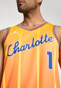 Oranžový basketbalový dres s žltými a modrými vertikálnymi pruhmi, na ktorom je v modrej kaligrafii napísané "Charlotte" a modré číslo "1". Textúrovaná látka.