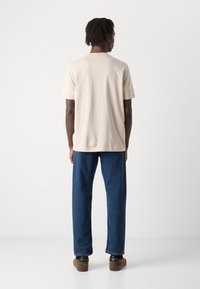 Lys beige bomulls t-skjorte med korte ermer, rett snitt. Kombinert med mørkeblå denimbukser, sett fra baksiden. Brune sko synlige.