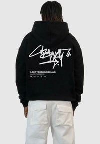 Črn kapucar z velikim belim grafičnim motivom in napisom na hrbtu, narejen iz mehkega materiala z ohlapnim krojem. Uporabljen z belimi hlačami.