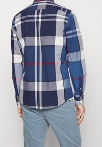 Man die een blauw-grijs geruit shirt met lange mouwen draagt, gecombineerd met lichtblauwe corduroy broek, gezien van achteren.