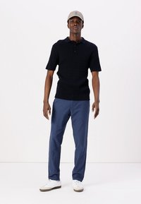 Mörkblå kortärmad pikétröja med texturerat mönster, kombinerad med blå byxor. Modellen bär en ljusgrå keps och vita sneakers.