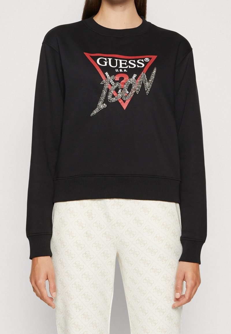 Sudadera negra con un diseño recortado que presenta un logo triangular rojo "GUESS U.S.A" y texto plateado brillante "ICON". Puños acanalados.