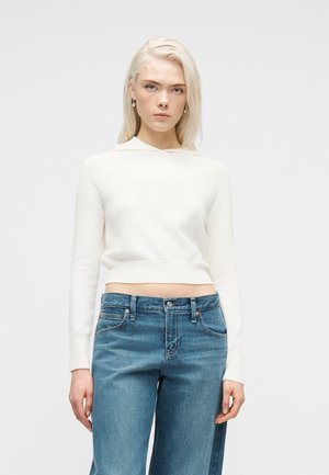 Młoda kobieta z blond włosami, nosząca srebrne kolczyki, biały krótki sweter z długim rękawem oraz wysokie niebieskie dżinsy, stojąca na tle szarego tła.