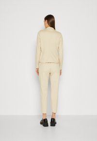 Veste beige ajustée et pantalon tapered assorti en tissu doux, présentant une texture lisse et un design minimal. Des chaussures noires complètent le look.