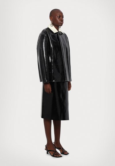 Giacca in pelle verniciata nera con maniche oversize, colletto bianco e gonna a tubino nera coordinata. Sandali neri con tacco e cinturini completano il look.