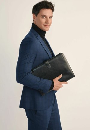 Czarna skórzana teczka trzymana przez osobę w granatowym, kratkowanym garniturze, z zamkiem błyskawicznym i uchwytem. Gładka tekstura.