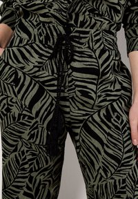 Green fabric pants with a black leaf pattern, featuring an elastic waistband and drawstring, pockets, and a relaxed fit.