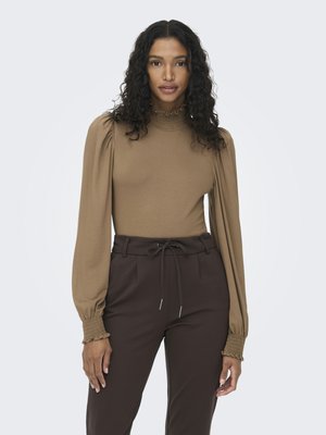 Mujer con cabello rizado que lleva una blusa beige de manga larga con hombreras abullonadas y pantalones de talle alto marrón oscuro con cintura ajustable.