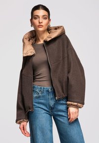 Femme portant une veste marron zippée avec doublure rembourrée beige, un chemisier taupe à manches longues, un jean bleu et des boucles d'oreilles créoles dorées, debout devant un fond clair.