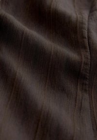Tmavě hnědá látka s jemnou vertikální žebrovanou strukturou a měkkými záhyby vytvářejícími jemné stíny.