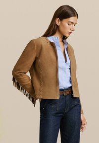 Kobieta w brązowej zamszowej kurtce z frędzlami, niebiesko-białej paskowanej koszuli, ciemnych dżinsach i brązowym pasku, stojąca na beżowym tle.
