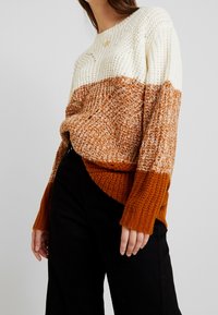 Sweter dzianinowy z dekoracyjnym zamkiem w kolorze kremowym, pomarańczowym i brązowym. Luźny krój z ściągaczami przy mankietach i u dołu, szczegóły z teksturowanej włóczki.