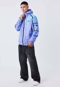 Giovane uomo che indossa una giacca a vento viola lucido con cappuccio, jeans neri larghi e sneaker bianche, in piedi contro uno sfondo chiaro e semplice.