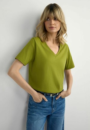 Kobieta o blond włosach, ubrana w oliwkową koszulkę z dekoltem w serek i niebieskie dżinsy, stoi z rękami w kieszeniach na tle białej ściany.