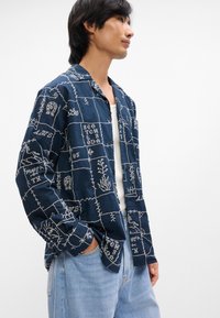 Ναυτικό μπλε πουκάμισο με λευκά κεντημένα σχέδια; διαθέτει κολάρο και κουμπιά, κατασκευασμένο από ελαφρύ ύφασμα.