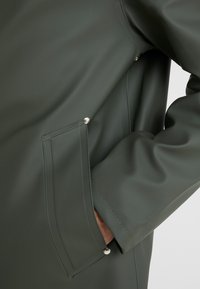 Osoba ubrana w ciemnozieloną wodoodporną kurtkę z ręką częściowo wsuniętą w boczną kieszeń z zatrzaskiem.