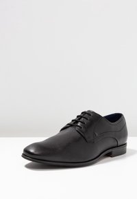 Chaussure noire en cuir texturé avec une finition lisse, présentant un design à lacets et un bout arrondi bas. Détail de marque discret.