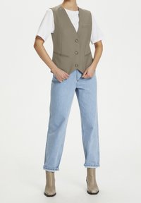 Kaki väst med djup V-ringning, tre knappar och framfickor, som bärs med ljusblå högmidjade jeans och beige ankelstövlar.