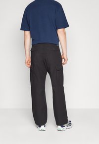 Person som bär svarta cargo-byxor med knäppta ficklock och vita sneakers, står mot en enkel ljus bakgrund.