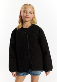 Sort quiltet jakke med en bølget tekstur, der har en rund halsudskæring og frontknaplukning, parret med denimshorts.