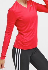 Rød langærmet atletisk top med V-hals og teksturerede ærmer, parret med sorte leggings, der har tre hvide striber ned ad siderne.