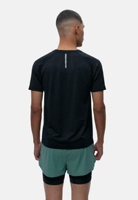 Juodas sportinis marškinėlis su tekstūruota medžiaga, trumpomis rankovėmis ir atspindinčia juostele nugarėlėje. Dėvimi su žaliais šortais.