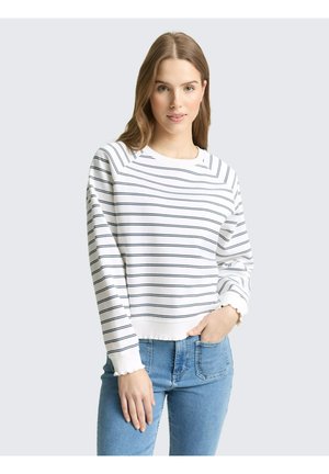 Młoda kobieta w białej bluzce z długim rękawem z cienkimi czarnymi paskami poziomymi i niebieskich dżinsach, stoi z jedną ręką w kieszeni.