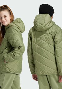 Giacca verde trapuntata con cappuccio. Presenta cuciture diagonali, tasche laterali e una silhoutte casual. Indossata con pantaloni verde chiaro.