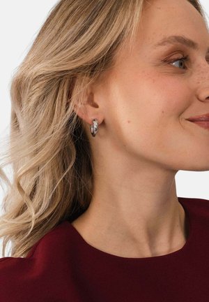 Blonde Frau mit welligem Haar, die silberne Creolen und ein burgunderrotes Oberteil trägt, lächelt und schaut nach rechts vor einem weißen Hintergrund.