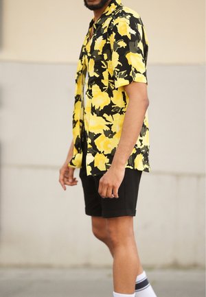 Mężczyzna w żółto-czarnej koszuli z krótkim rękawem w kwiaty, czarnych szortach, białych skarpetkach, stojący na utwardzonej powierzchni na tle beżowej ściany.