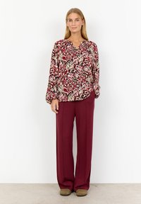 Röd och krämfärgad mönstrad blus med lös passform, kombinerad med vida vinröda byxor. Bruna slip-ons fulländar outfiten.