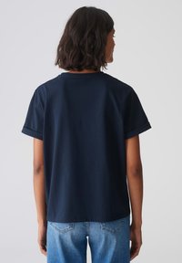 T-shirt in cotone blu navy con maniche corte, con risvolti sui polsini e una vestibilità rilassata, vista da dietro su uno sfondo bianco semplice.