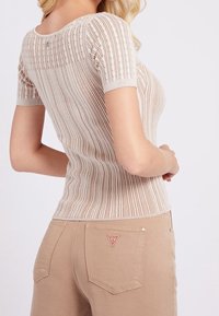 Mujer vistiendo una camiseta de manga corta color beige con relieve y pantalones color tostado con el logo de GUESS en el bolsillo trasero, mostrada desde el lado y la parte trasera.
