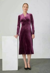Samta, ar garām piedurknēm, purpura kleita ar pieguļošu augšdaļu, V veida dekoltē un ceļgala garuma svārkiem. Modelis valkā melnas kurpes pret neitrālu fonu.