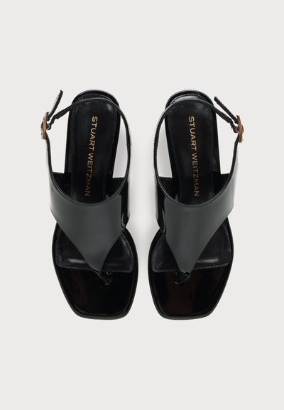 Μαύρα σανδάλια slingback από δερμάτινο λουστρίνι με λουράκια ανάμεσα στα δάχτυλα και χρυσλό λογότυπο "Stuart Weitzman" στις εσωτερικές σόλες, εμφανιζόμενα από πάνω σε απλό φόντο.