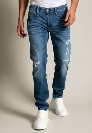 Homme portant un jean bleu usé et des baskets blanches, debout devant un fond neutre.