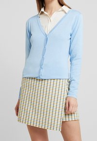 Světle modrý cardigan vyrobený z měkkého materiálu, s výstřihem do V, dlouhými rukávy a zapínáním na knoflíky. V kombinaci se sukní s kostkovaným vzorem v žluté a modré barvě.