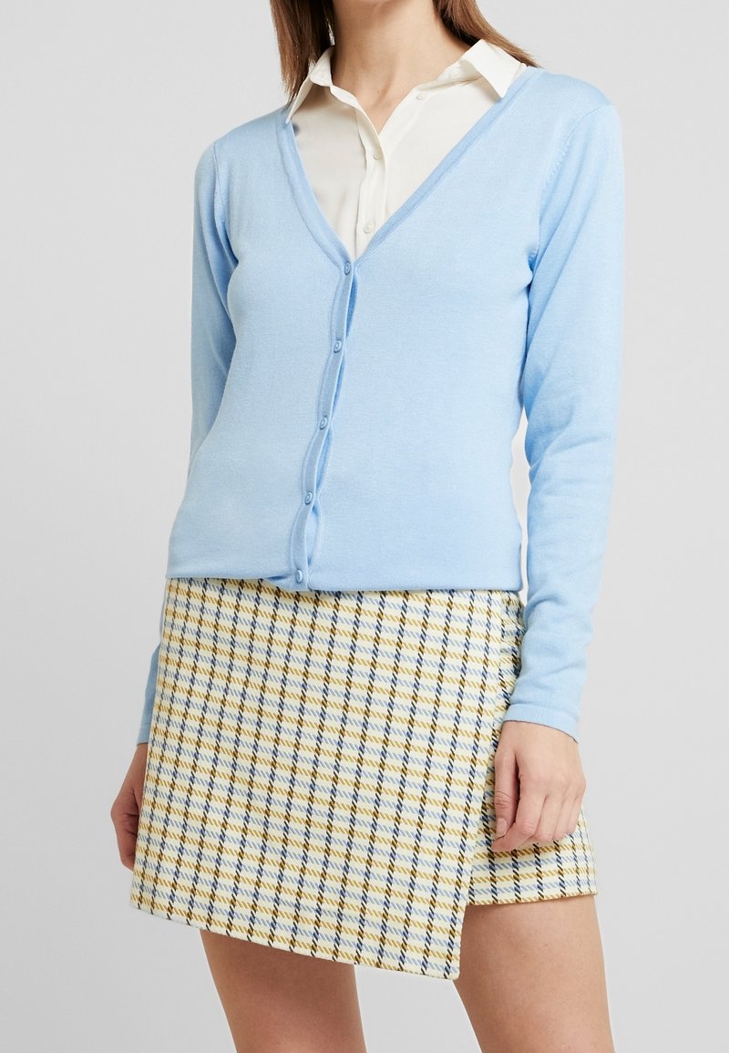 Světle modrý cardigan vyrobený z měkkého materiálu, s výstřihem do V, dlouhými rukávy a zapínáním na knoflíky. V kombinaci se sukní s kostkovaným vzorem v žluté a modré barvě.