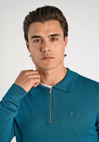 Tealfärgad pikétröja med dragkedja, gjord av ett mjukt, strukturerat tyg; har en krage och en broderad logga på vänster bröst.