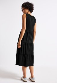Robe sans manches noire avec des volants superposés au niveau de la jupe, fabriquée en tissu doux. La vue arrière montre une encolure arrondie et un détail de logo.