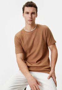 Brun tekstureret T-shirt med korte ærmer og hvidt halsudskæring, parret med lyse bukser. Model siddende og vendt fremad.