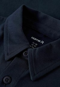 Polo shirt blu navy con colletto classico, due bottoni neri e una superficie in cotone morbido. L'etichetta mostra la taglia EU 92, taglia US 2T.