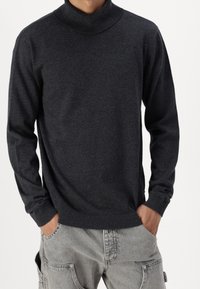 Uomo che indossa un maglione a collo alto grigio scuro e pantaloni cargo grigio chiaro con le mani in tasca, su uno sfondo neutro.