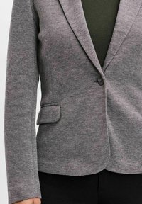 Šedý vzorovaný blazer s klopovým límcem, zapínáním na jeden knoflík a klopovou kapsou. Tonální textura, přiléhavý střih, nošený přes tmavě zelený top.