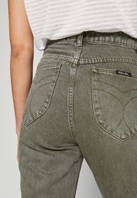 Jeans de mezclilla verde oliva con un diseño de cintura alta, que cuentan con bolsillos traseros con un patrón de costura curva distintivo y etiqueta de marca.